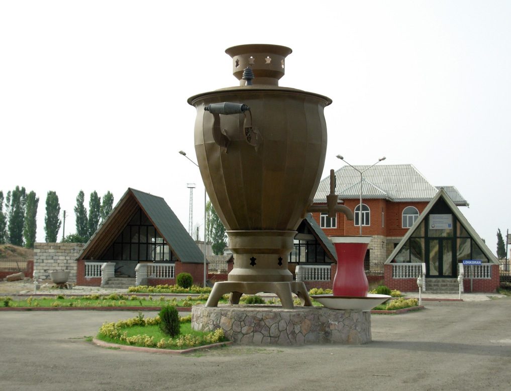 A giant samovar monument with a tea glass beside it, in a landscaped garden in Lankaran