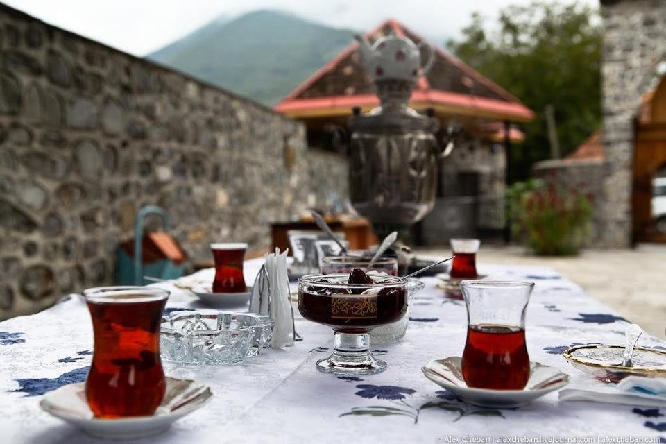 Glasses of tea and a bowl of jam on a table outdoors with a samovar and mountains behind