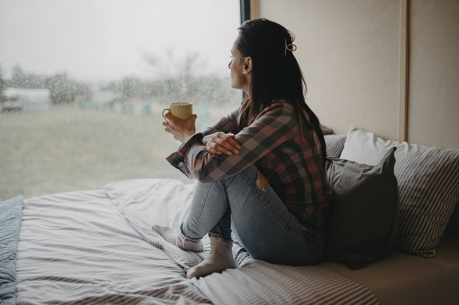 A woman sitting on a window seat with a hot drink, watching the rain outside