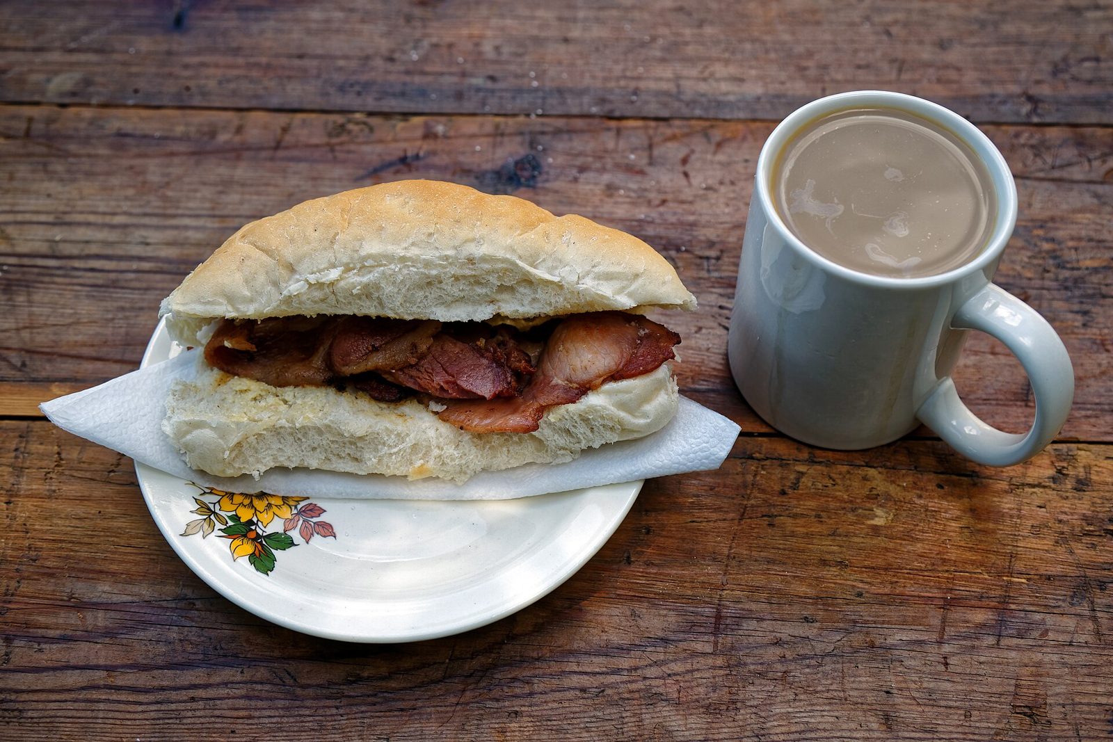 A bacon roll on a plate next to a mug of milky tea on a wooden table