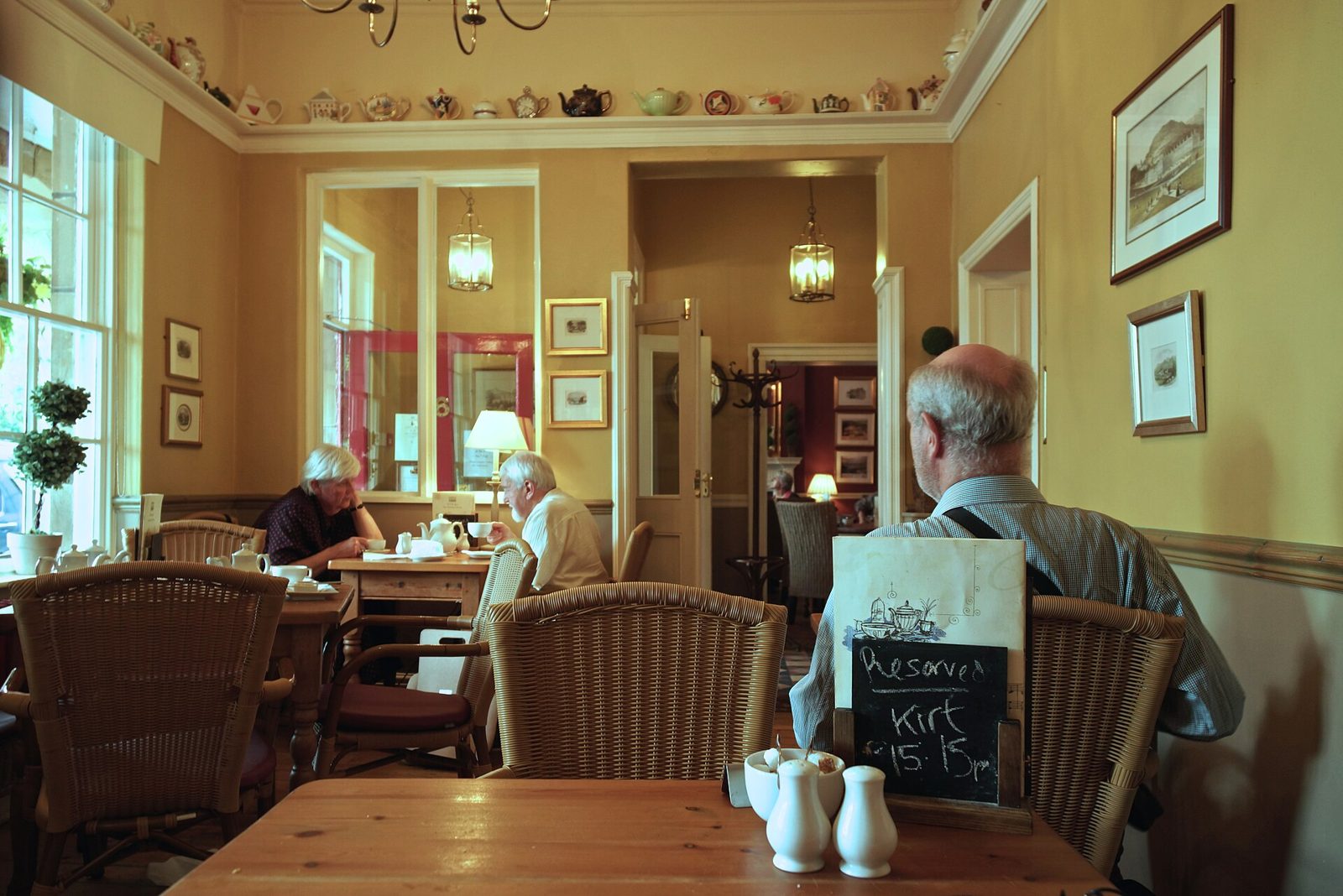Customers sitting at wooden tables in a traditional English tea room with teapots on a high shelf